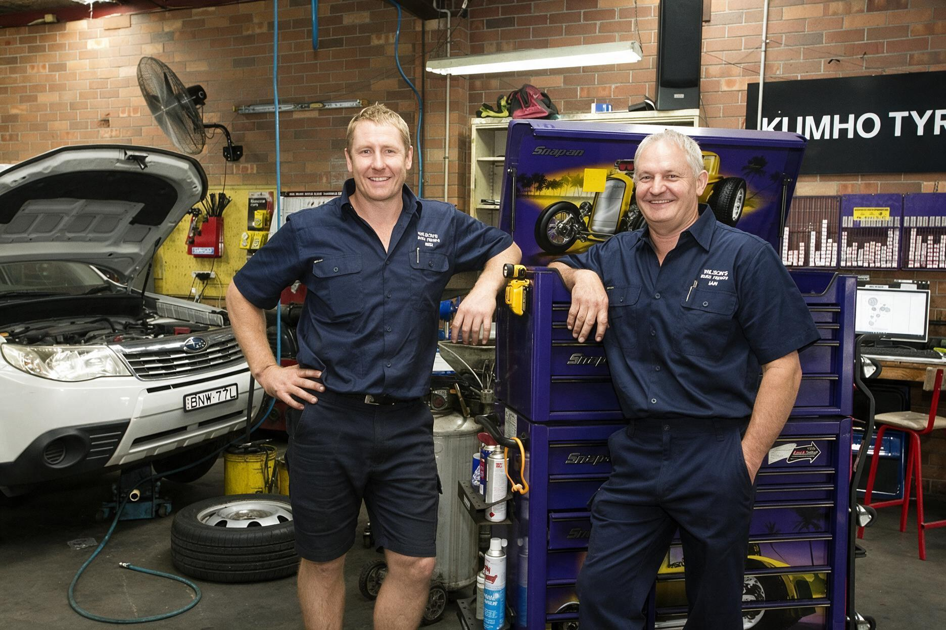 Two mechanics in navy uniforms standing in an auto repair shop beside a lifted car and tool cabinet