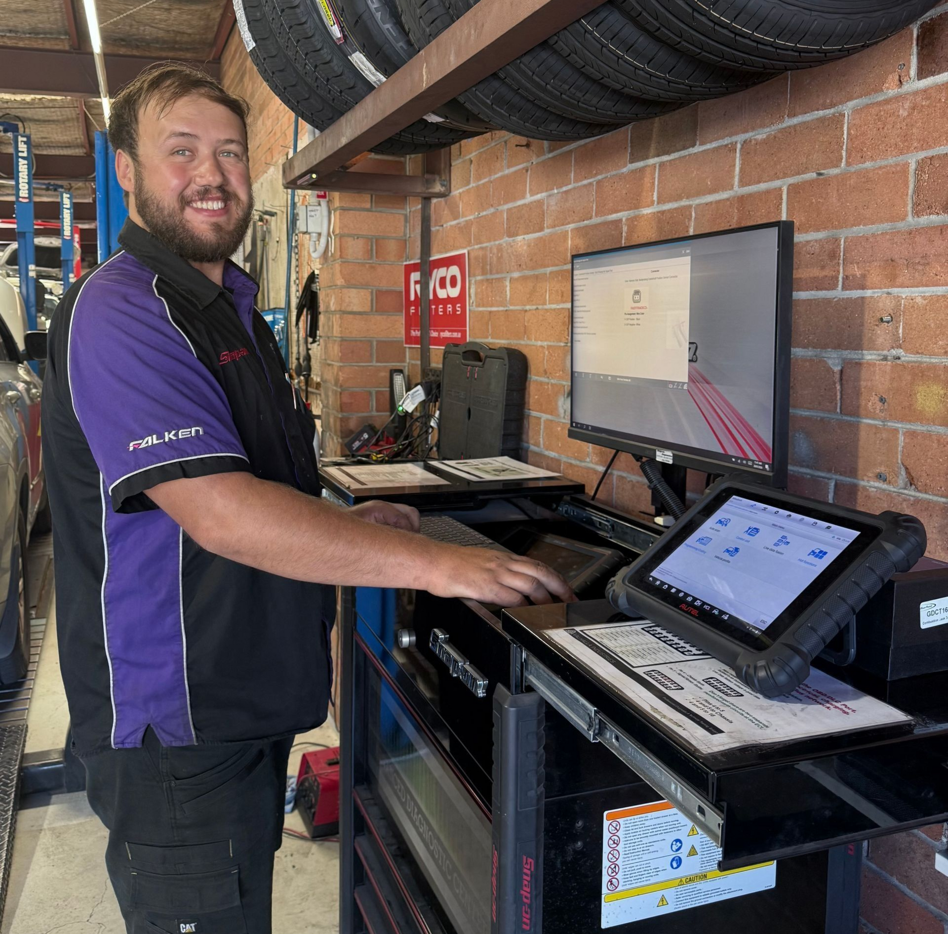 Mechanic smiling at a workbench with a computer monitor and tablet in an auto shop
