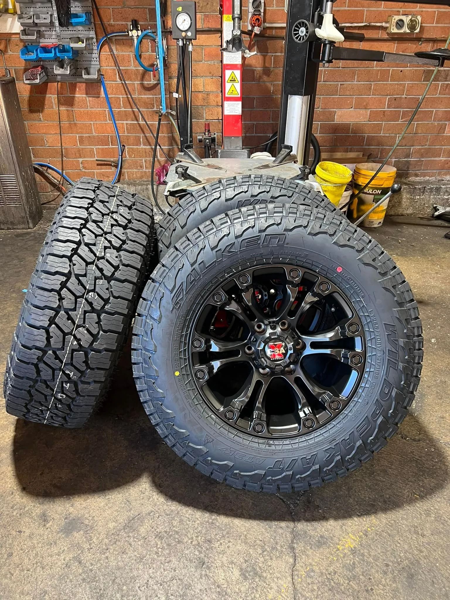 Two off-road tires with black rims leaning against a brick wall in a workshop.