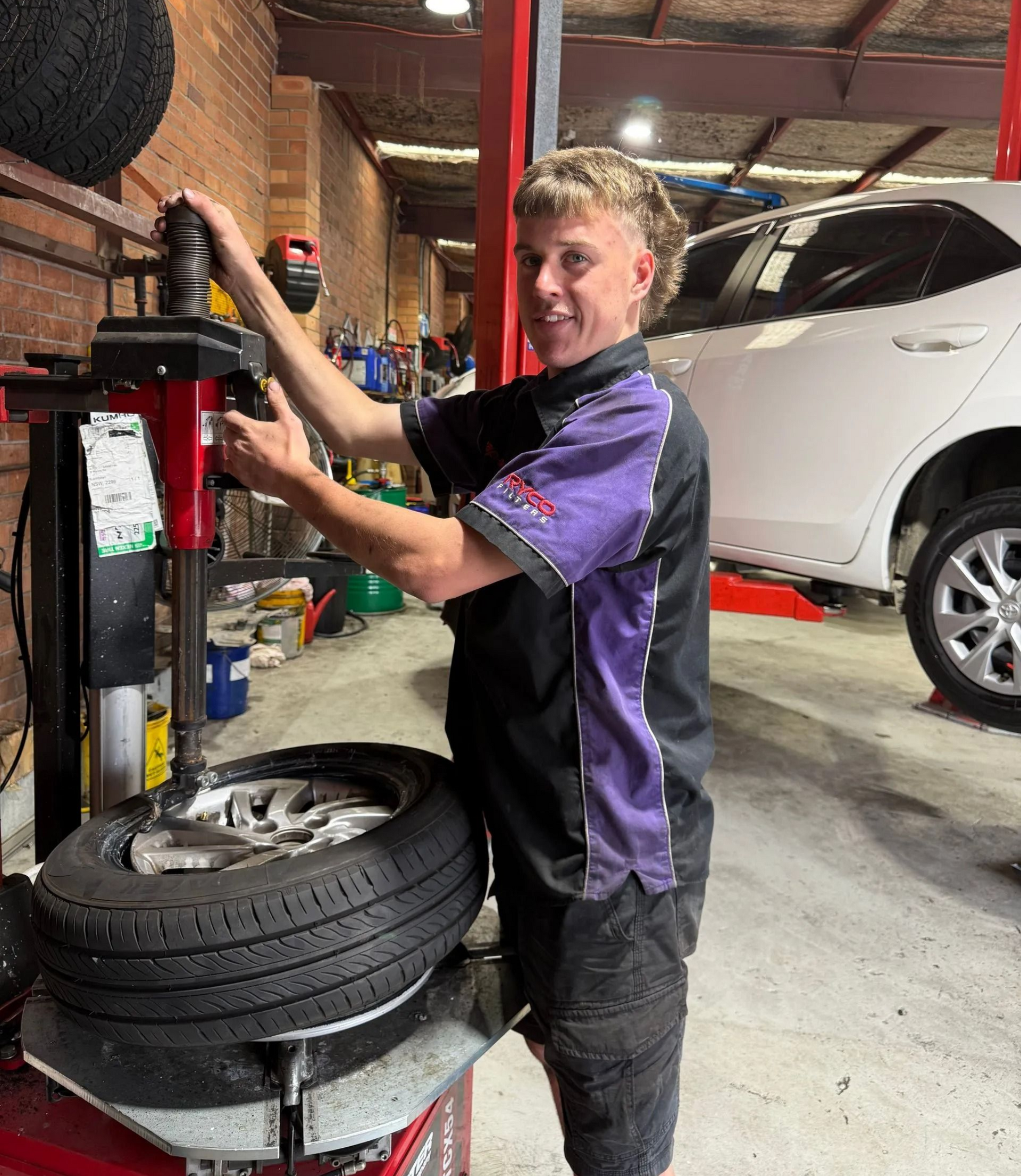 Mechanic using a tire-changing machine in an auto shop, with a white SUV in the background.