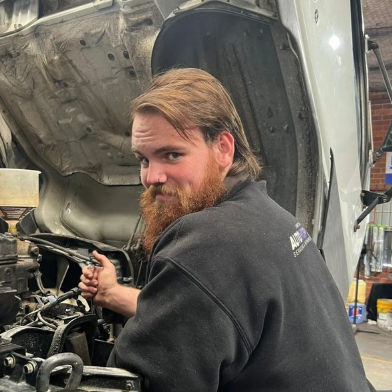 Mechanic working under a car’s open hood in an auto shop, looking back at the camera.