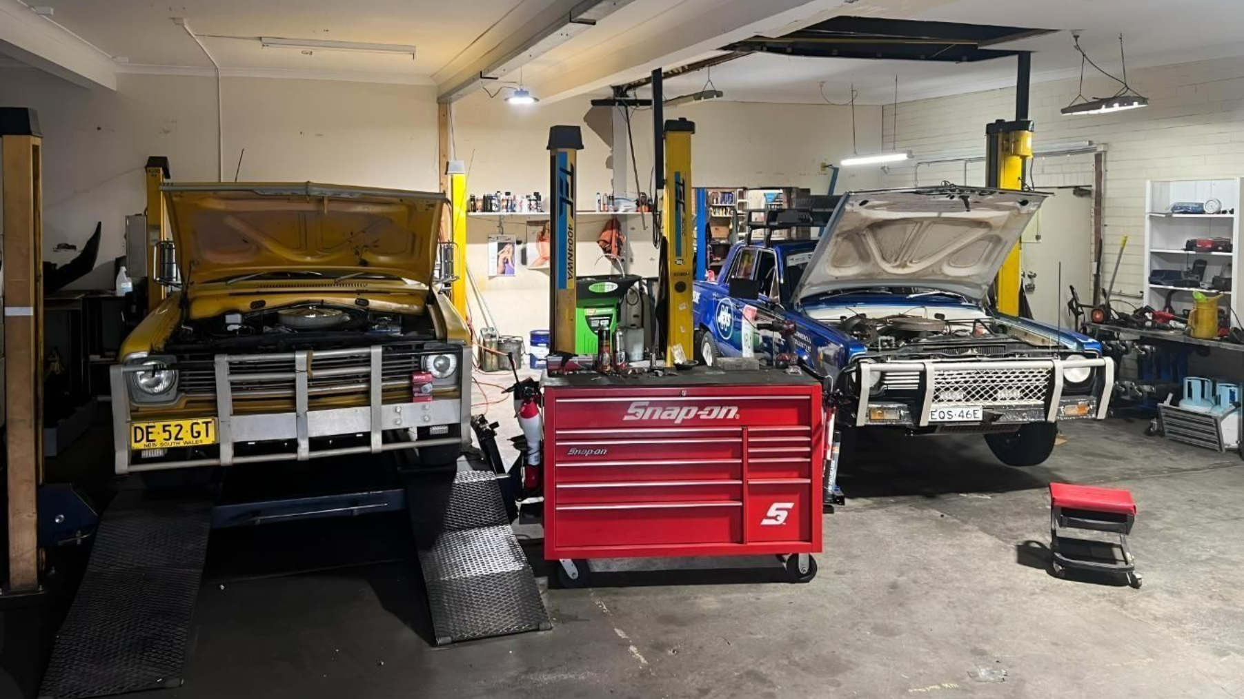 Two vehicles under repair in a garage, with a red tool chest centered between them.