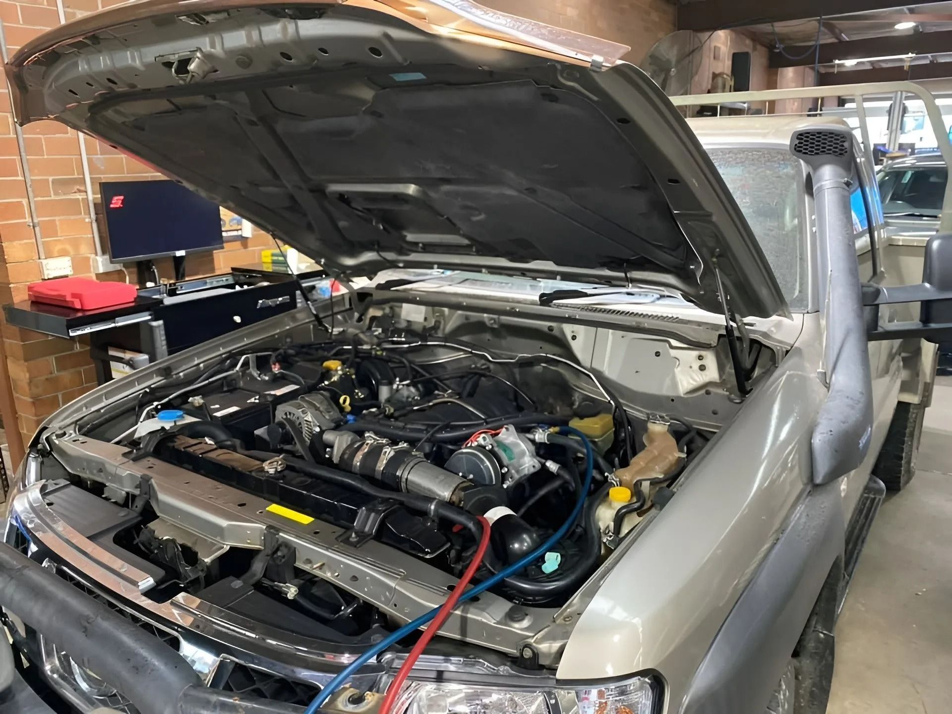 Mechanic working under the hood of a silver car beside an air-conditioning service machine in a garage