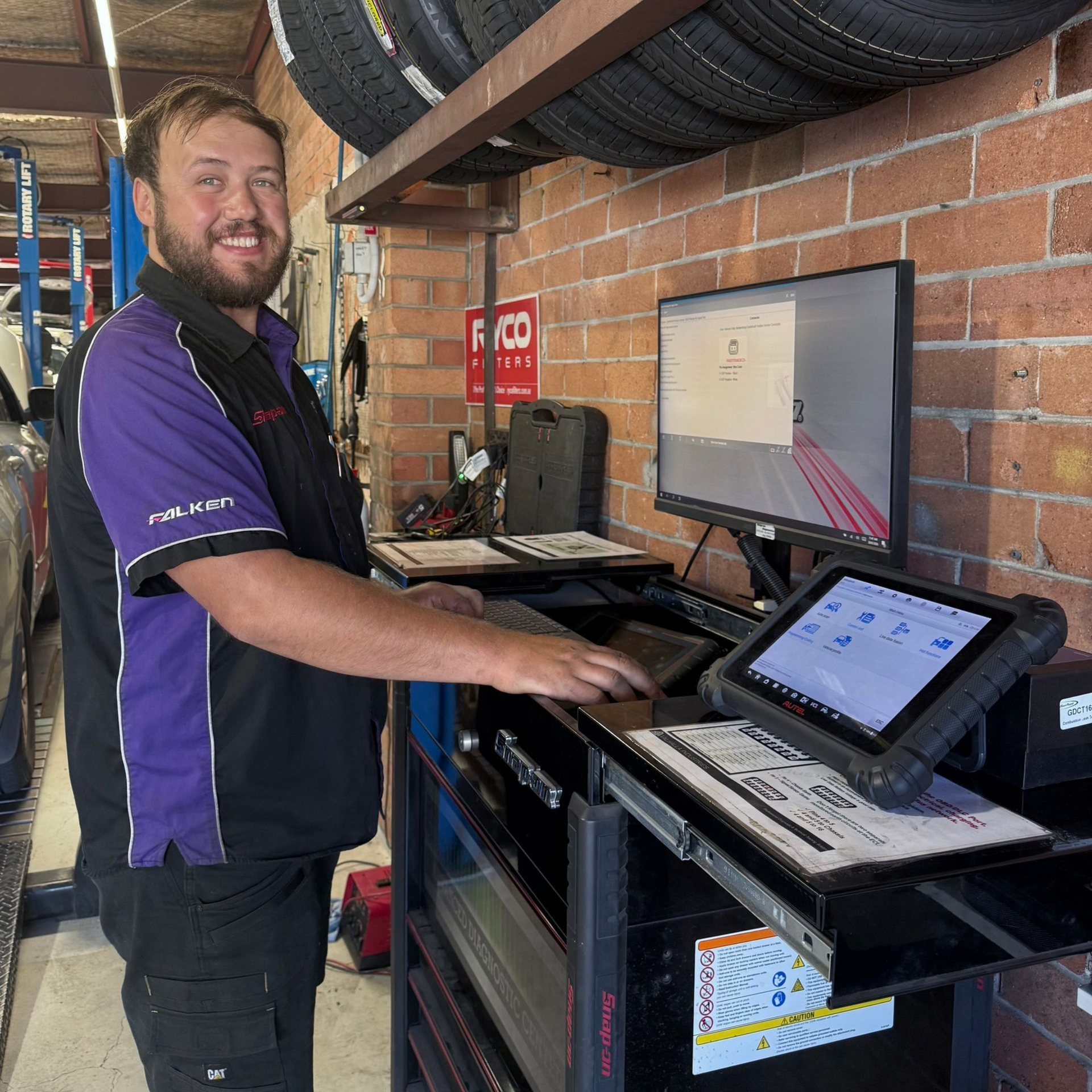Mechanic using a diagnostic computer at a garage workbench, with tires stored on shelves behind him.