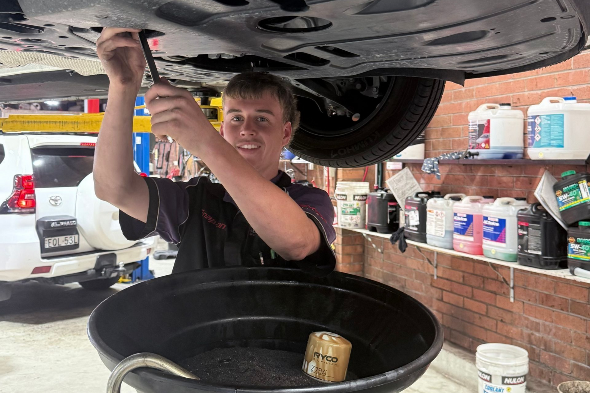 Mechanic draining oil under a lifted car in a garage, with a large catch pan below.