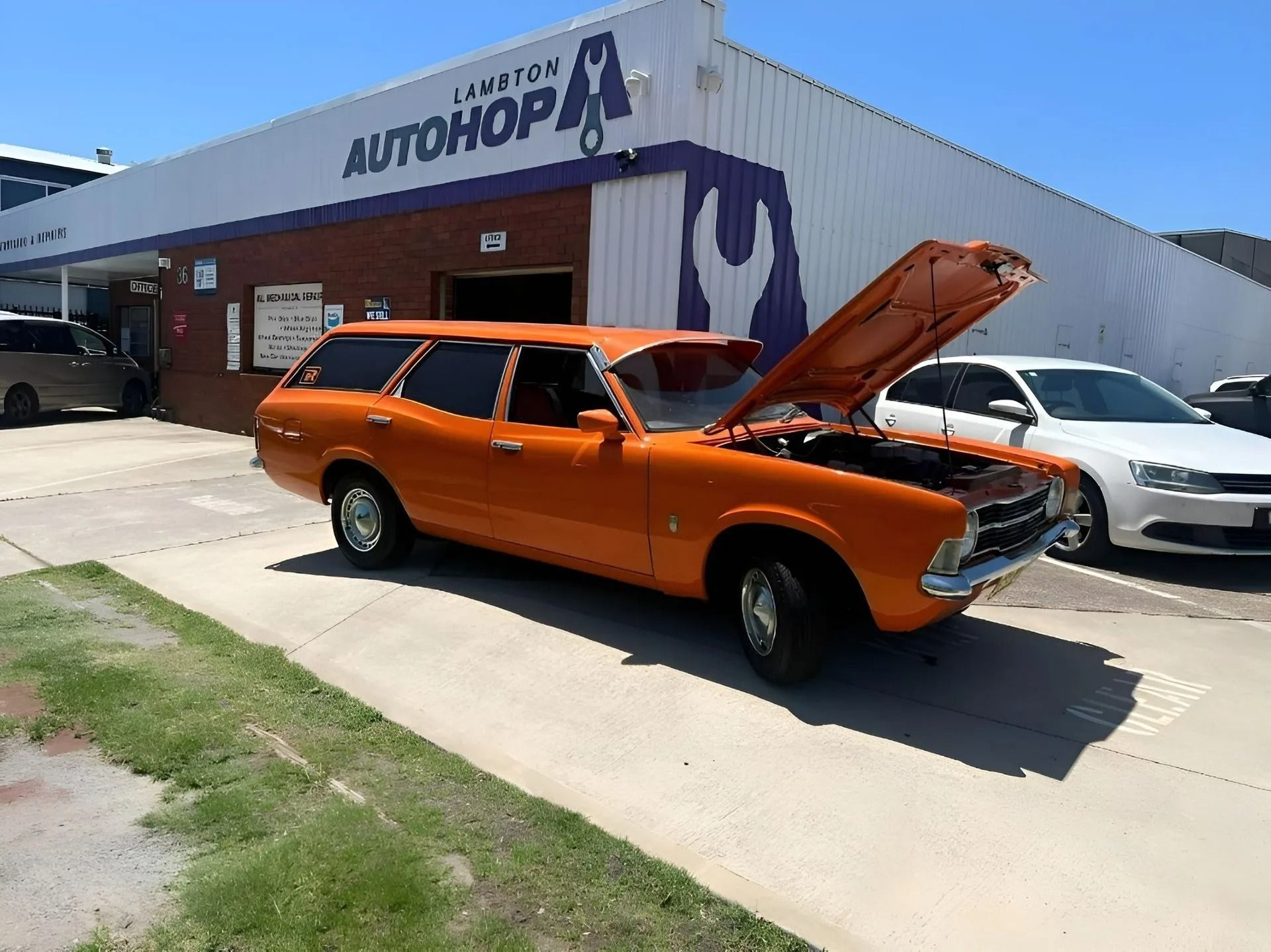 Orange classic car with hood open parked outside an auto shop on a sunny day