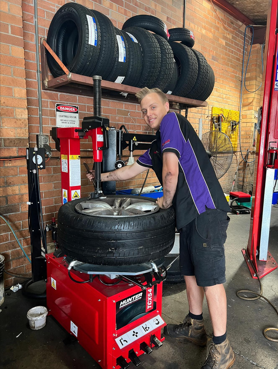 Mechanic changing a tire on a red tire machine in a workshop with stacked tires.