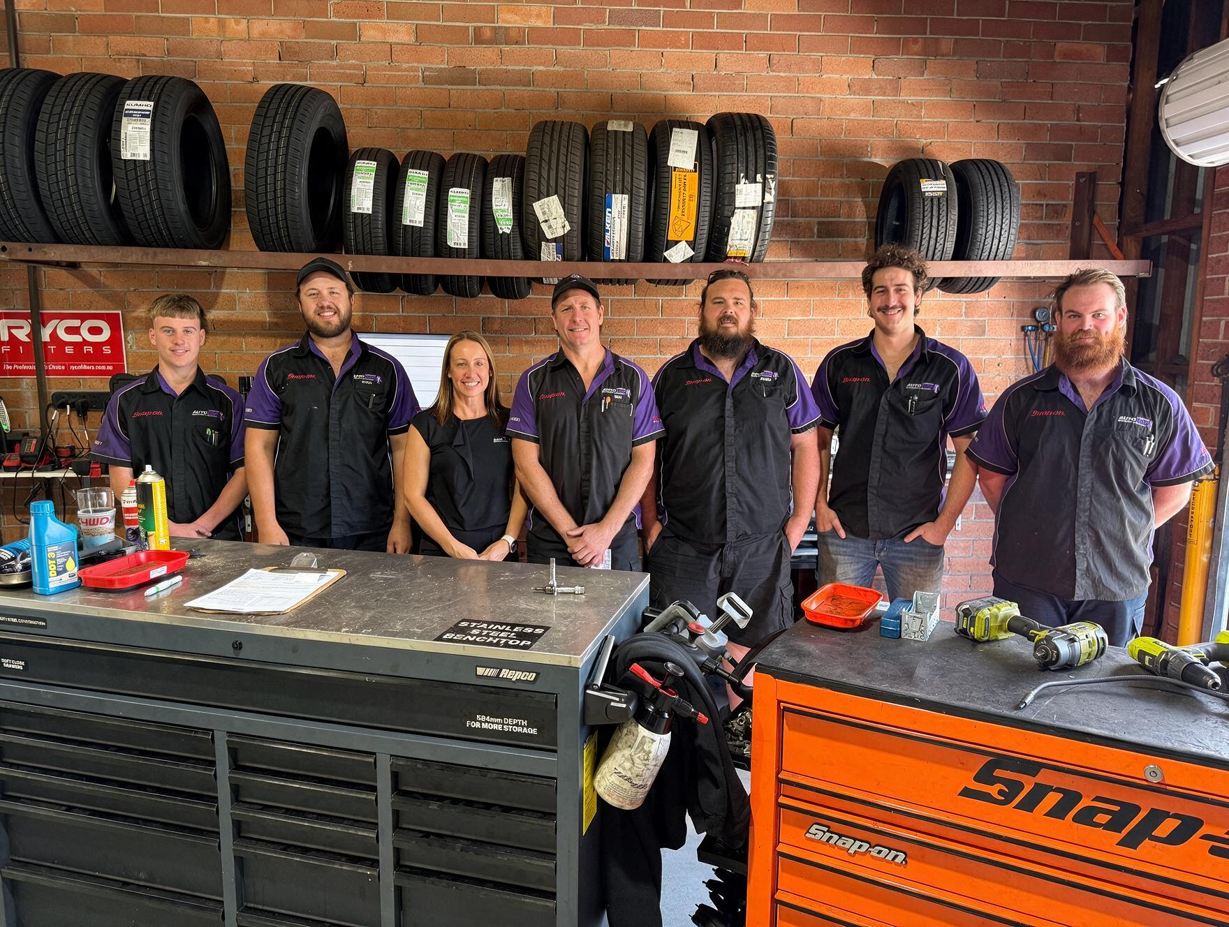 Six mechanics standing in a tire shop beside tool chests and stacked tires