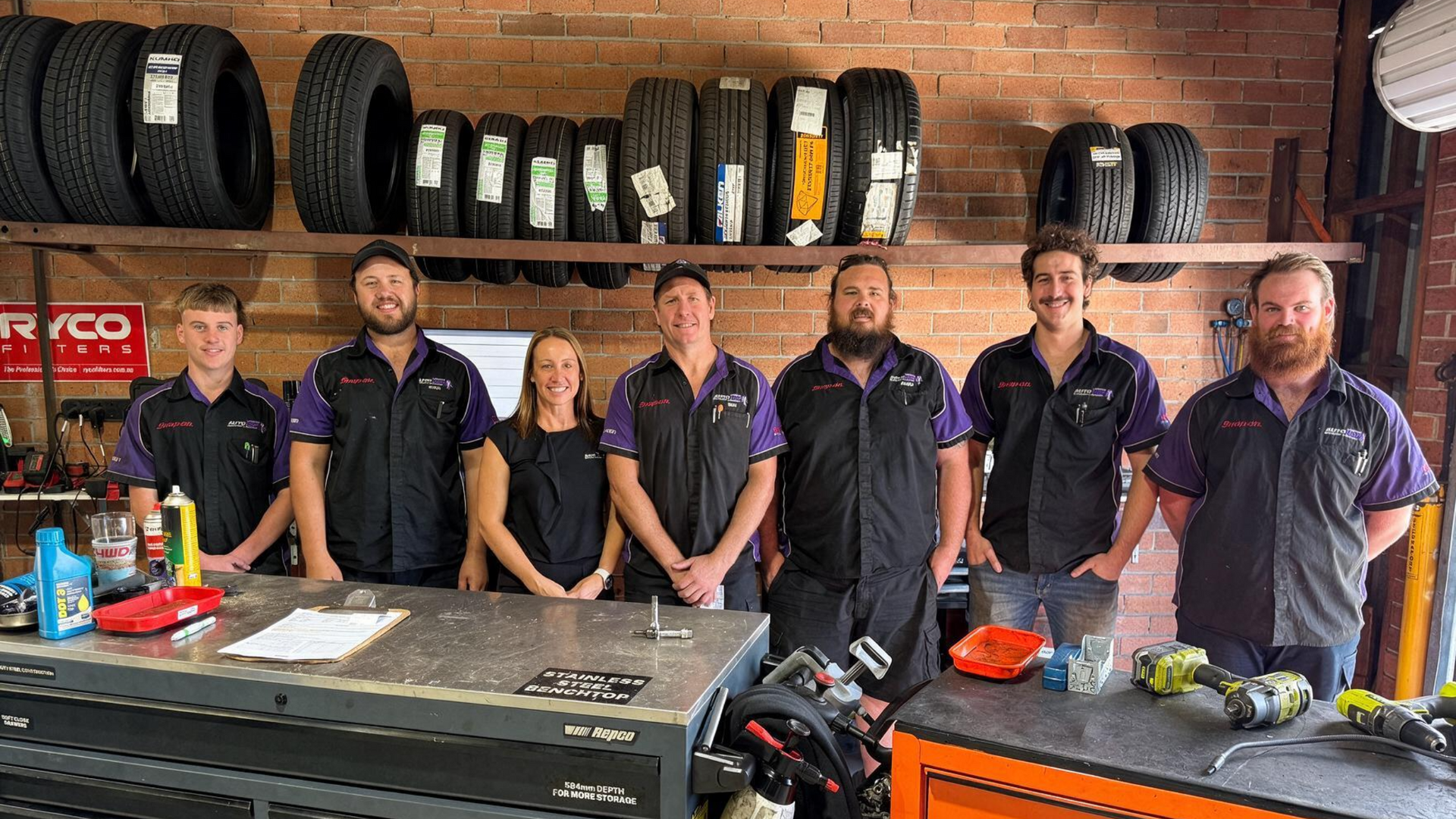 Six mechanics standing behind a workbench in an auto shop with tires stacked on shelves.