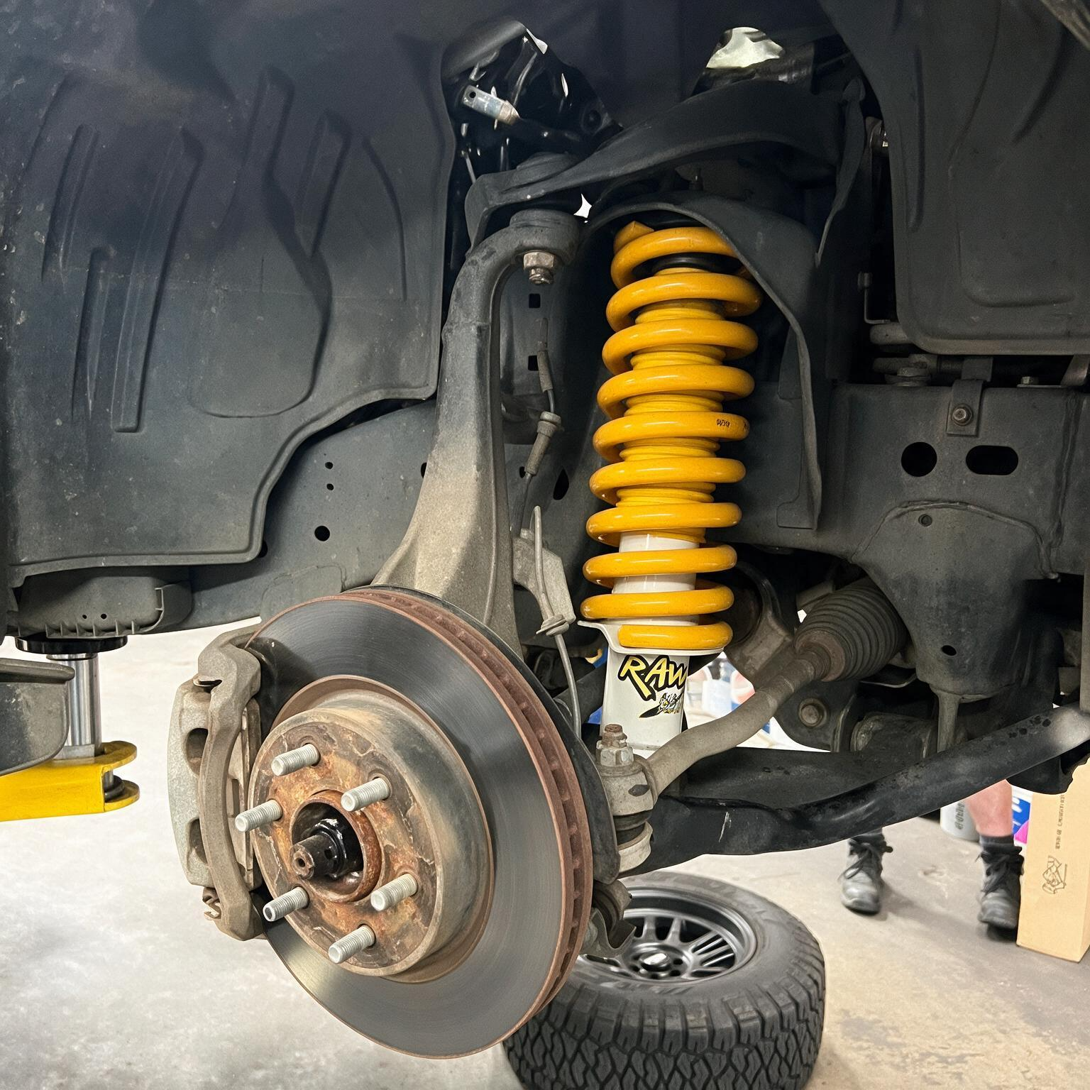 Car wheel removed, exposing brake rotor and yellow suspension spring in a garage.