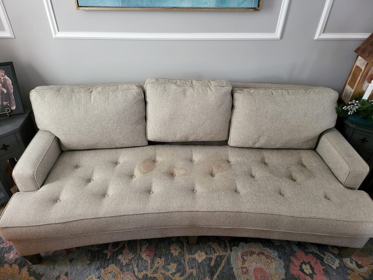 Beige curved sofa with three back cushions and button detailing, stained in the center.