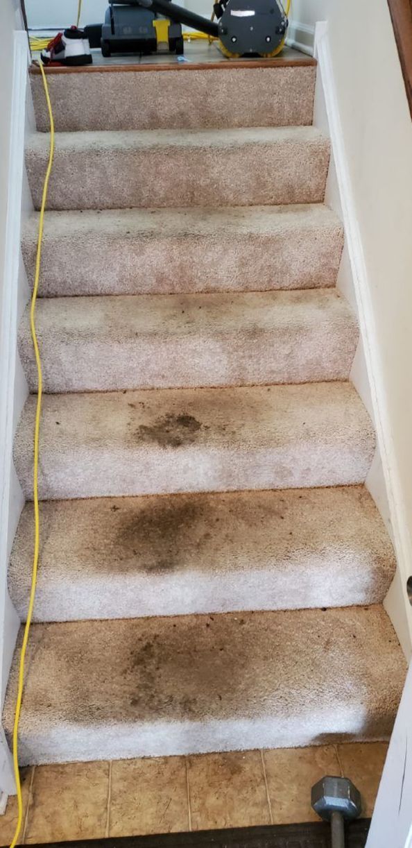 Dirty, light-colored carpeted stairs, with dark stains, are ready to be cleaned.  A cleaning machine rests at the top.