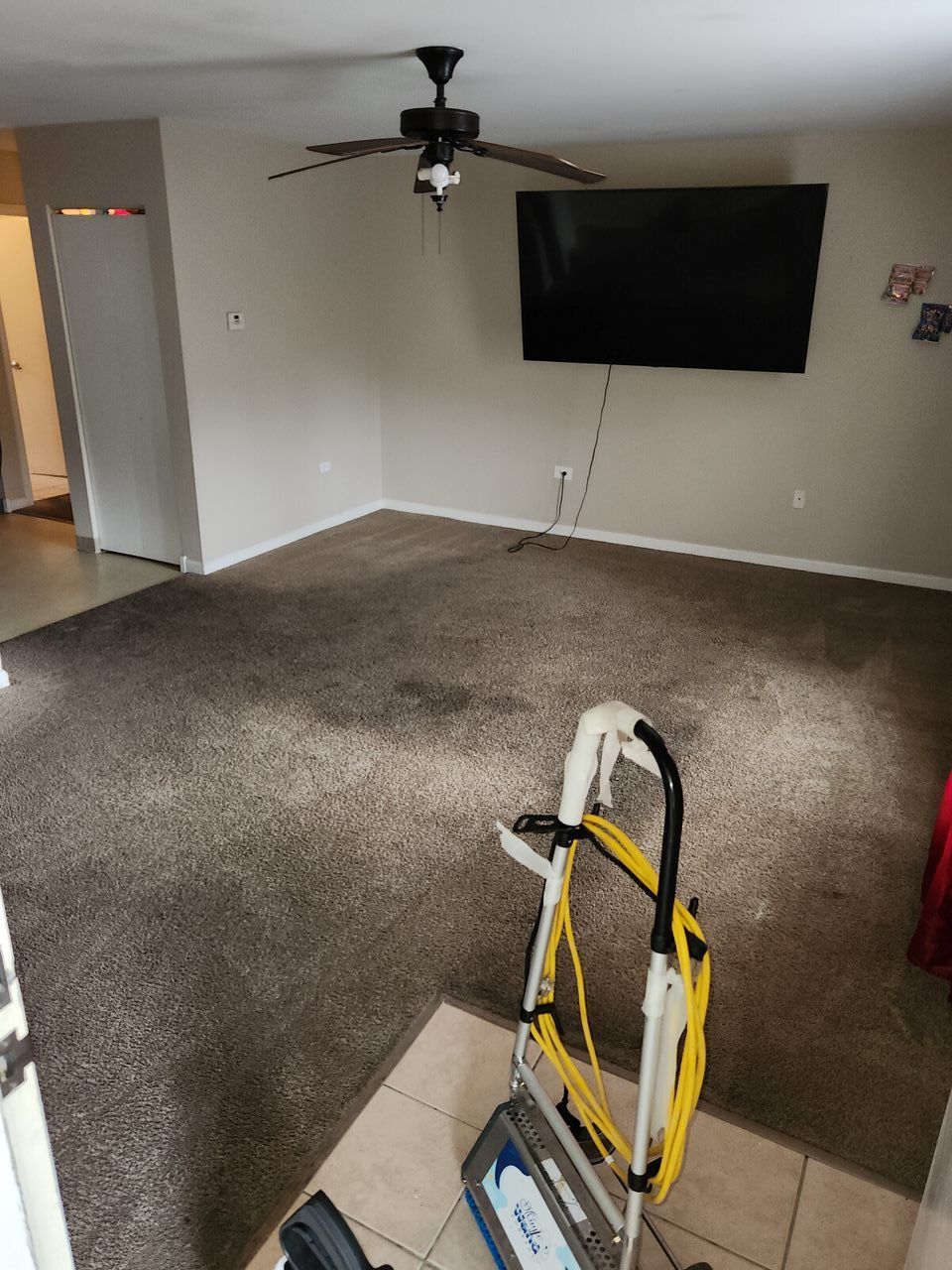 Living room with brown carpet, TV on wall, ceiling fan, and cleaning equipment.