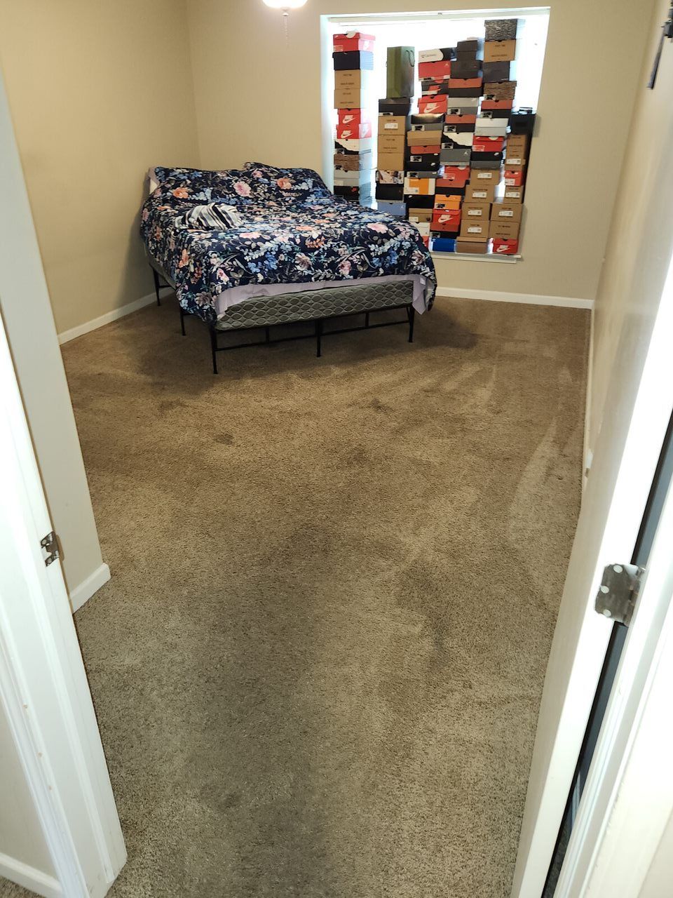 Bedroom with bed, shoe boxes stacked in window. Beige carpet, light-colored walls.