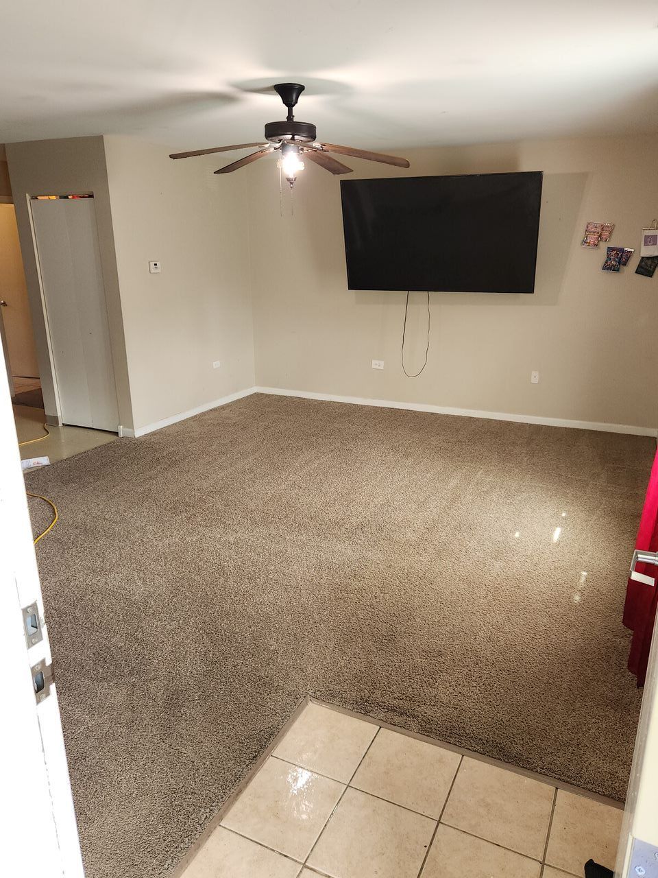 Living room with brown carpet, tan walls, ceiling fan, TV, and open doorway.