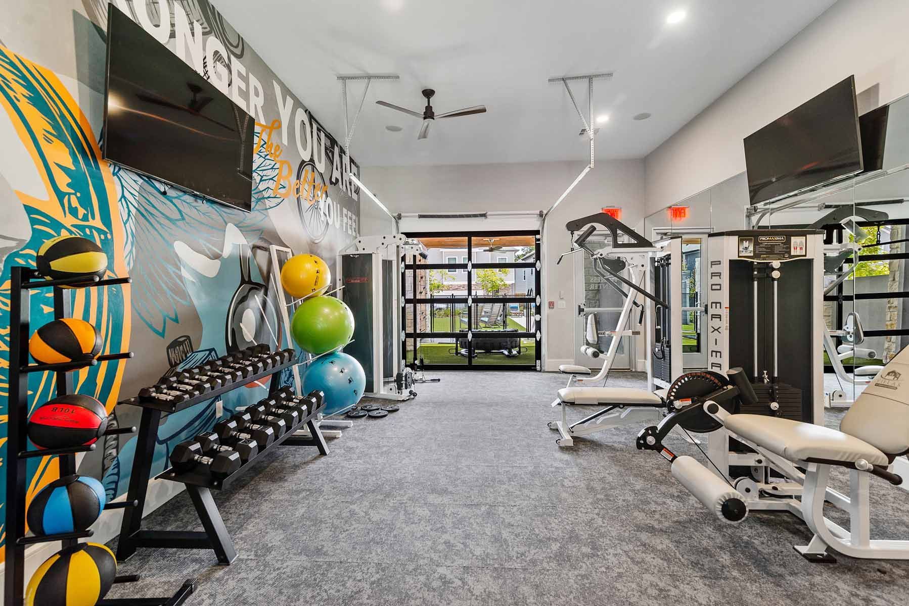 Gym with weights, exercise machines, and colorful wall art. Open doorway leads to outdoor view.