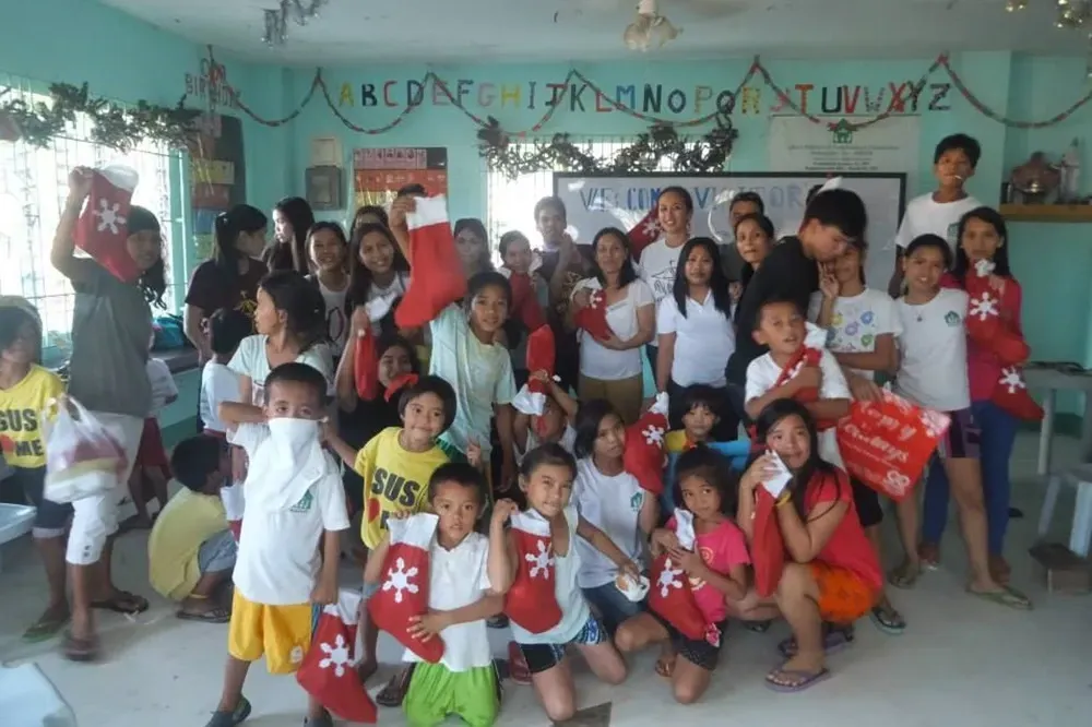 Group of children and adults posing indoors with red gift bags in a decorated classroom