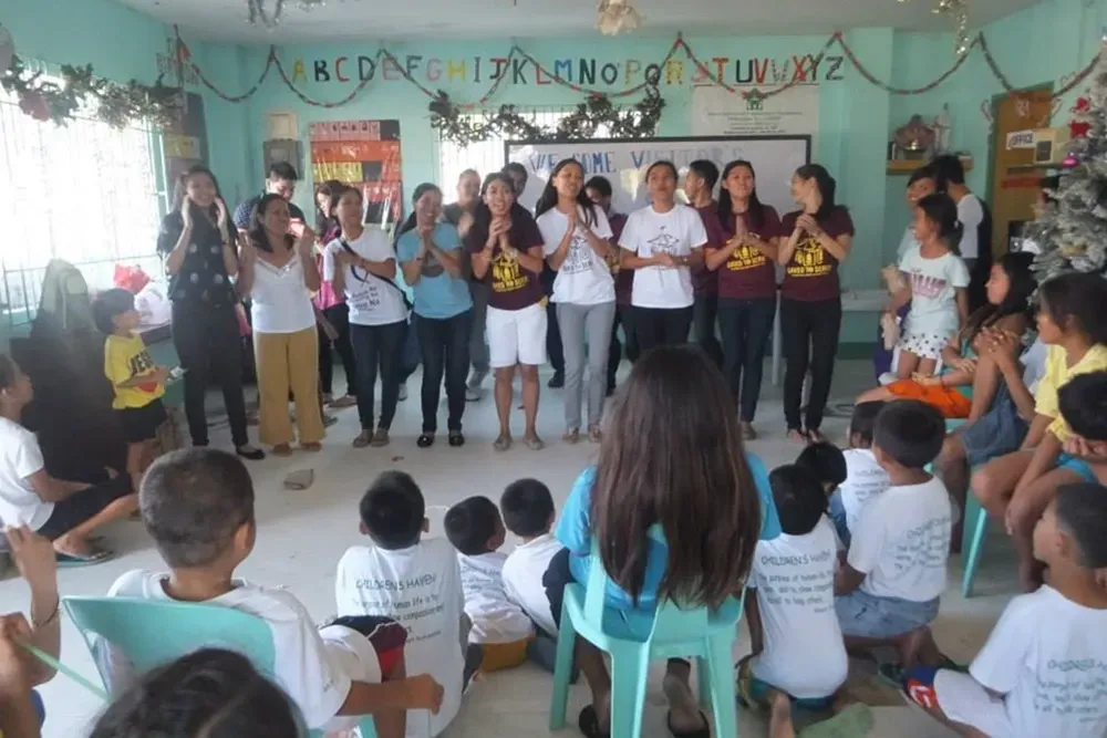 Group of people standing and clapping in a decorated classroom, with children seated and watching.