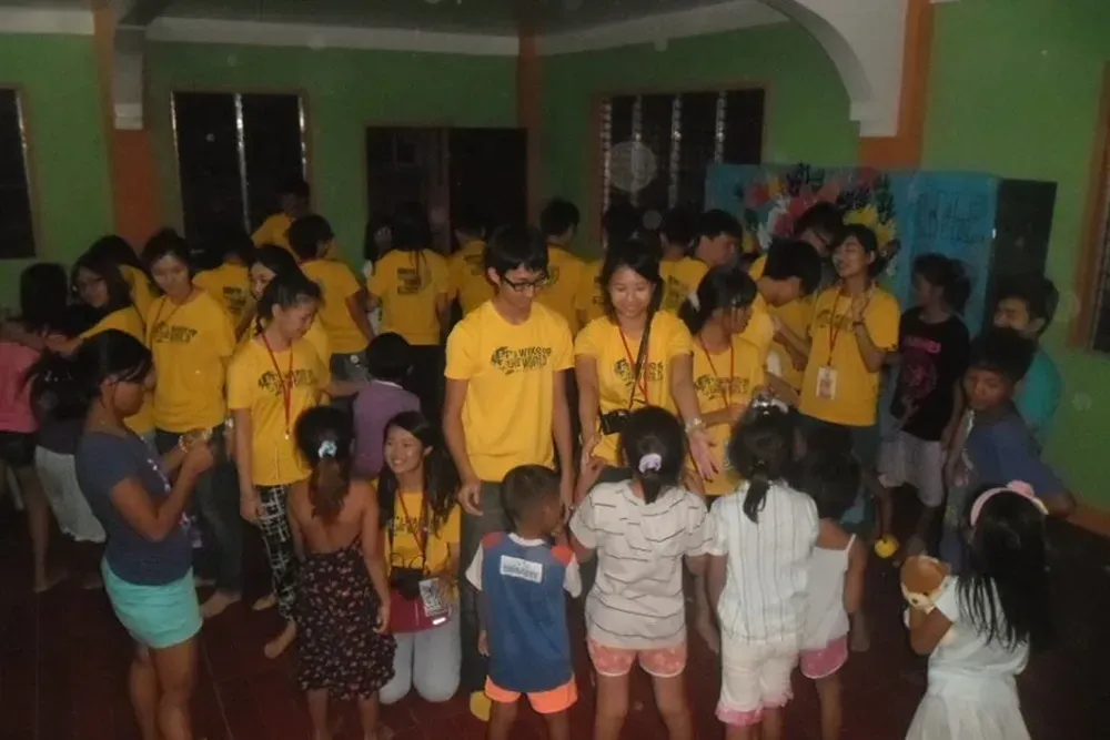 Group gathered indoors, many in yellow shirts, listening to a speaker in a dimly lit room.