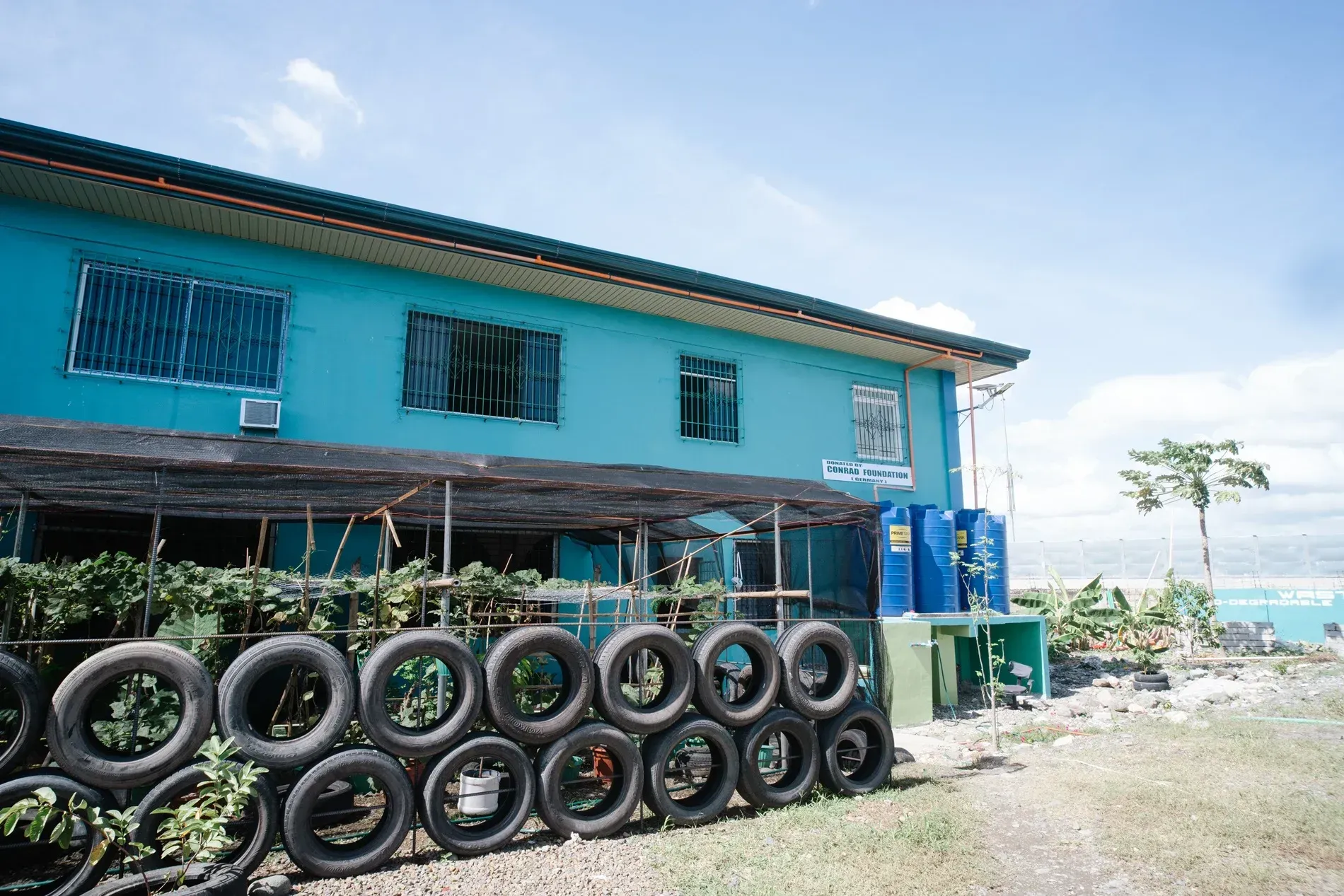 Blue waterfront building with stacked tires along the front and a gravel road beside the sea