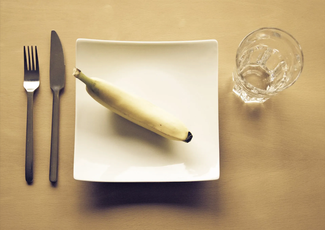 A banana on a white plate with silverware and a glass of water, set on a wooden surface.