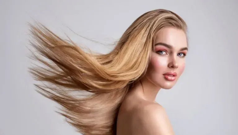 Blonde woman with flowing hair, rosy makeup, and bare shoulders poses against a gray background.