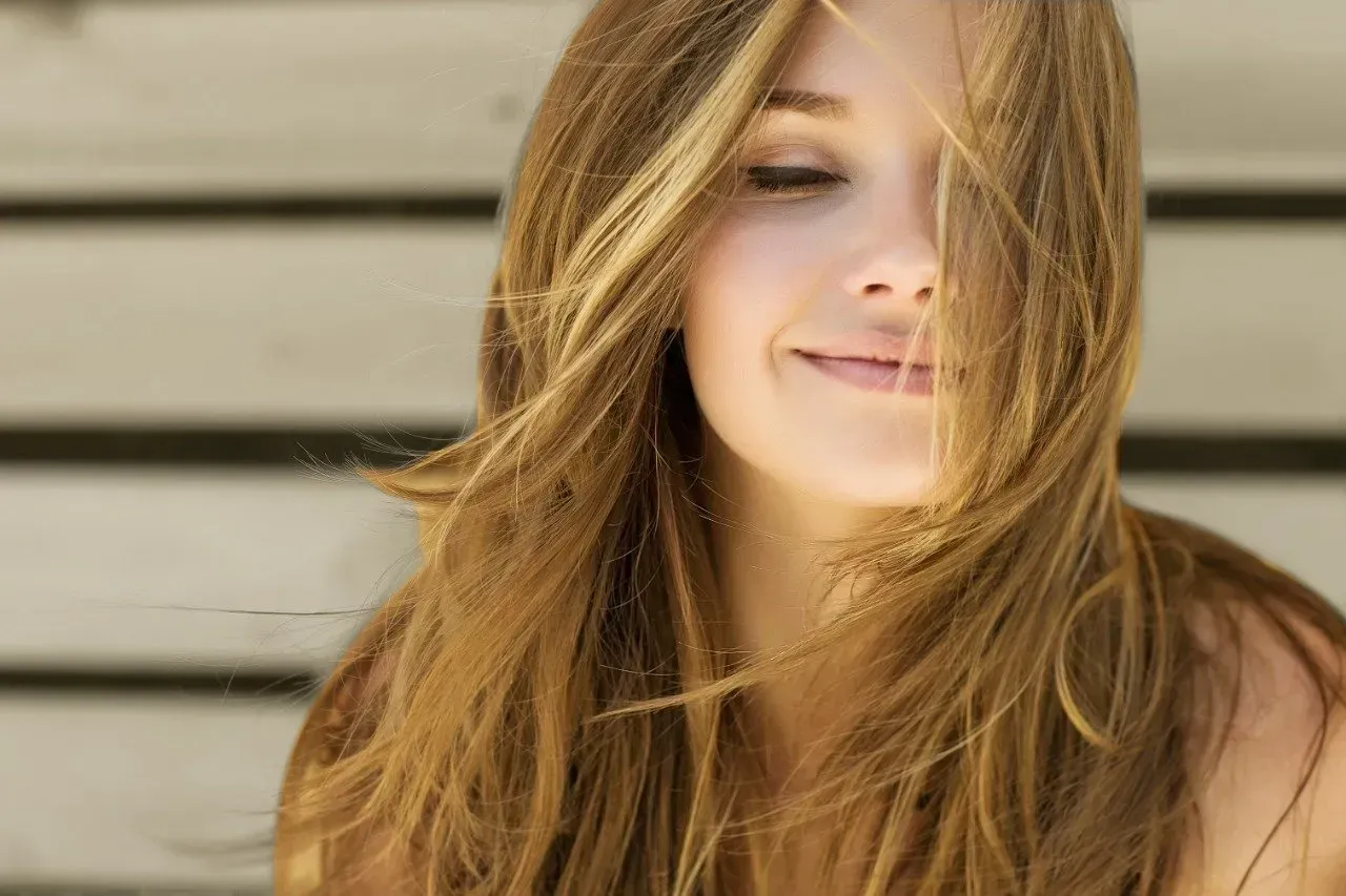 Woman with long, light brown hair smiling gently in front of a light-colored wooden wall.