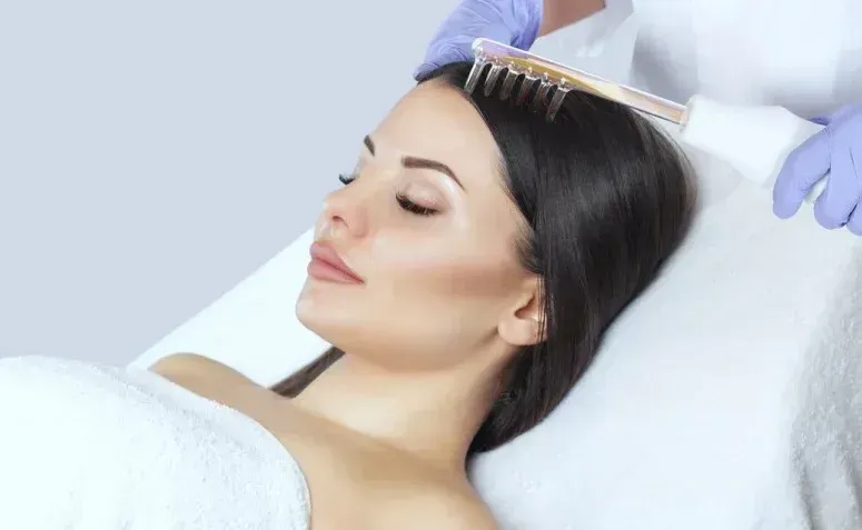 Woman receiving scalp treatment with a comb-like device. A gloved hand holds the device on her head while she reclines, eyes closed, in a medical setting.