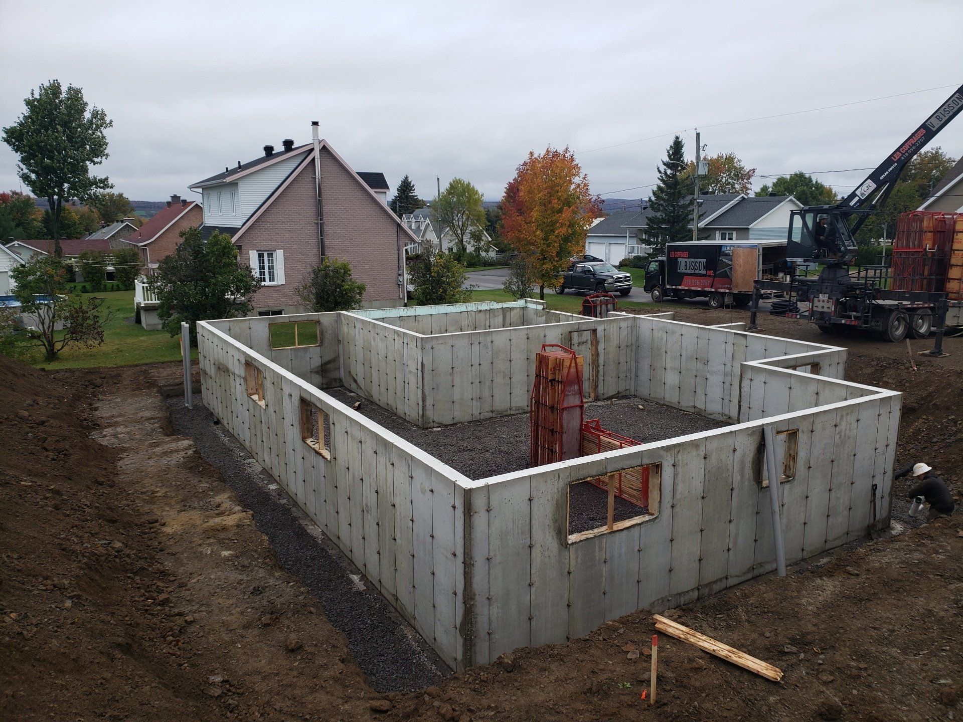 Fondations en béton d'une maison en construction sur un terrain résidentiel. Murs gris, remblai de gravier et petites fenêtres visibles.