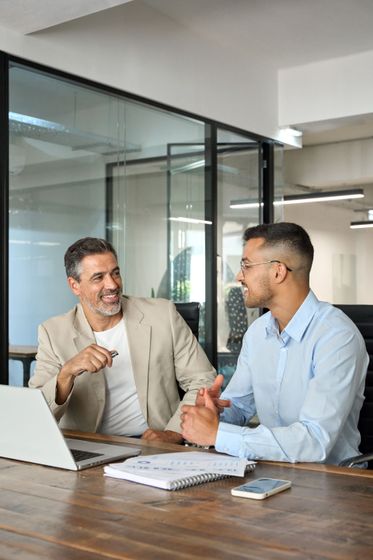 Two colleagues discussing at a desk in a modern office with a laptop and notebooks.
