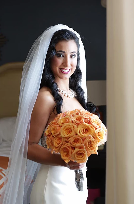 Bride in white veil holding a bouquet of orange roses, smiling indoors.