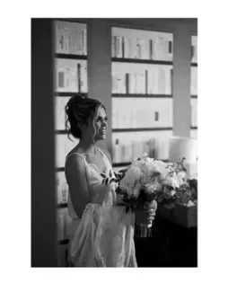 Woman holding a bouquet in a room with shelves and a window, standing in profile and smiling.