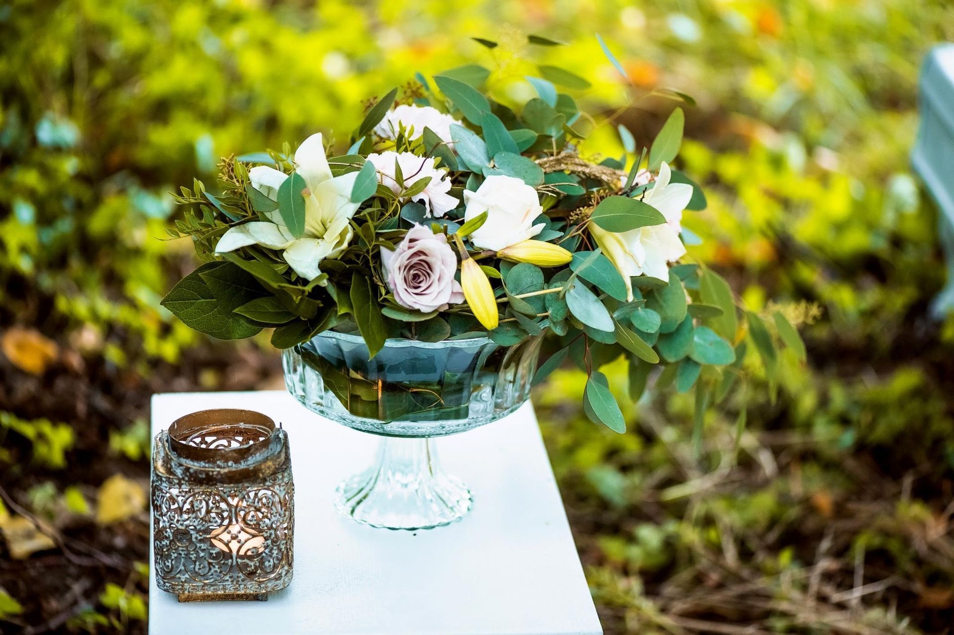 White floral bouquet in a glass vase on a table outdoors with a candle lantern, against a green garden background