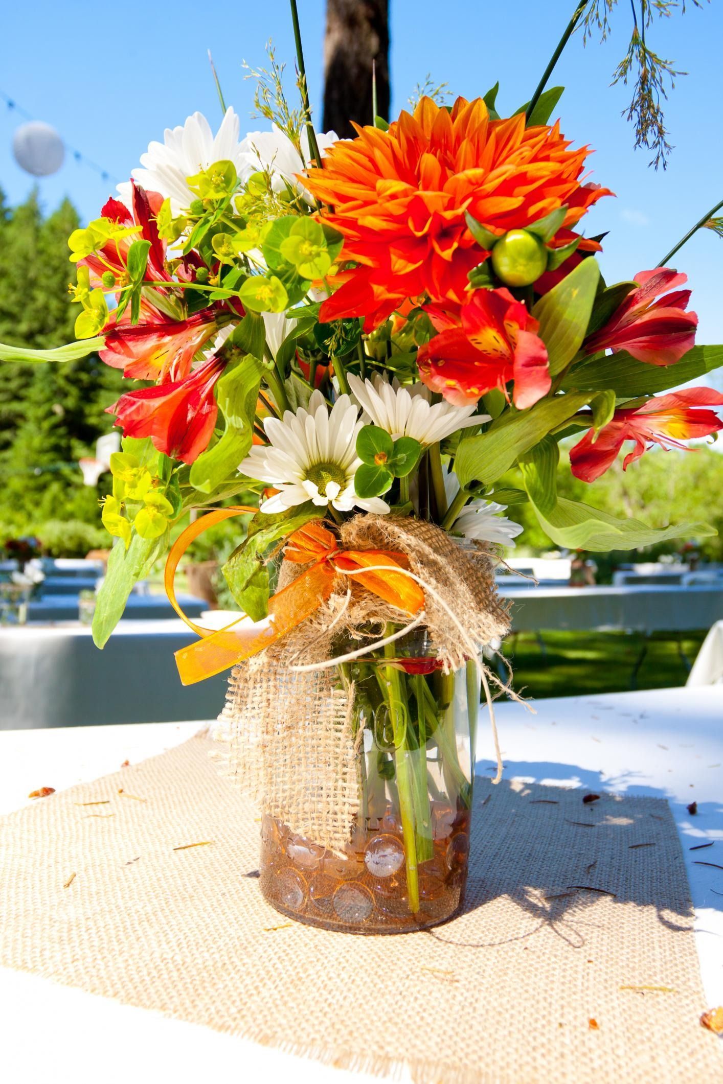 Colorful mixed bouquet of orange, red, and white flowers in a glass vase wrapped with burlap outdoors