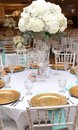 Elegant banquet table with white floral centerpiece, gold chargers, and white chairs in a softly lit reception hall