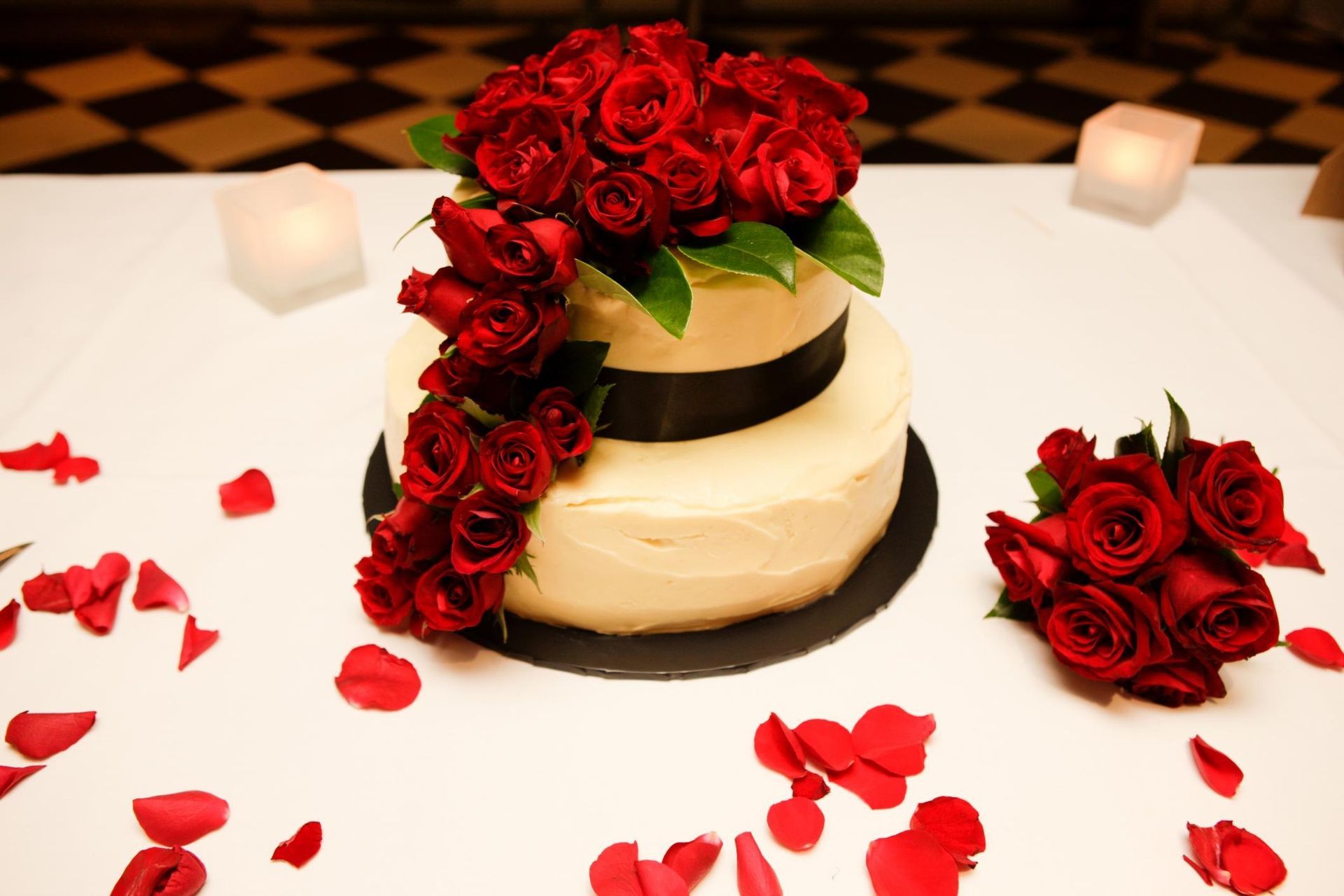 Red roses on a tiered cake with scattered petals on a table, set among soft candlelight.
