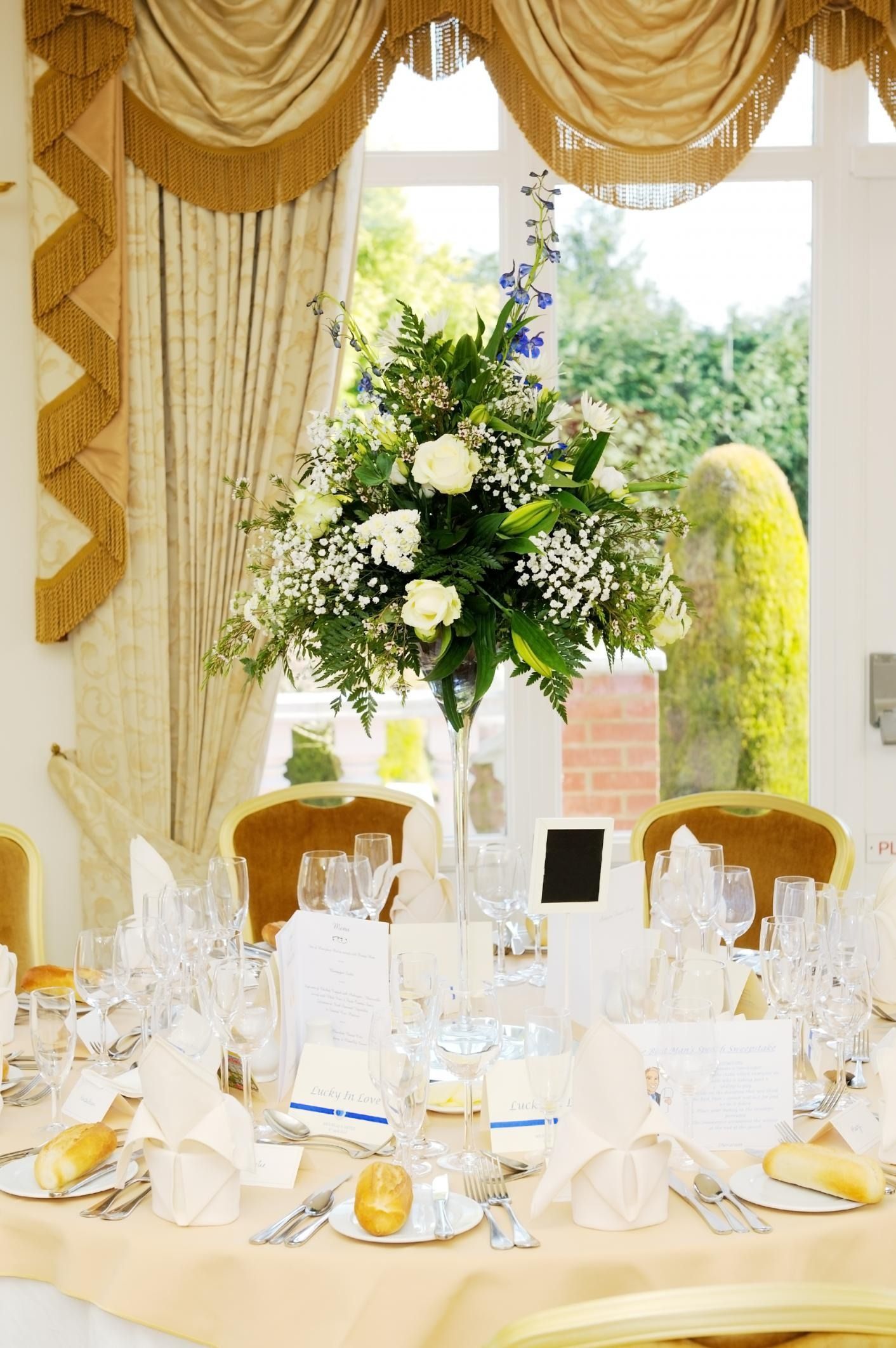 Elegant dining table set by a sunlit window with gold drapes and a tall white floral centerpiece