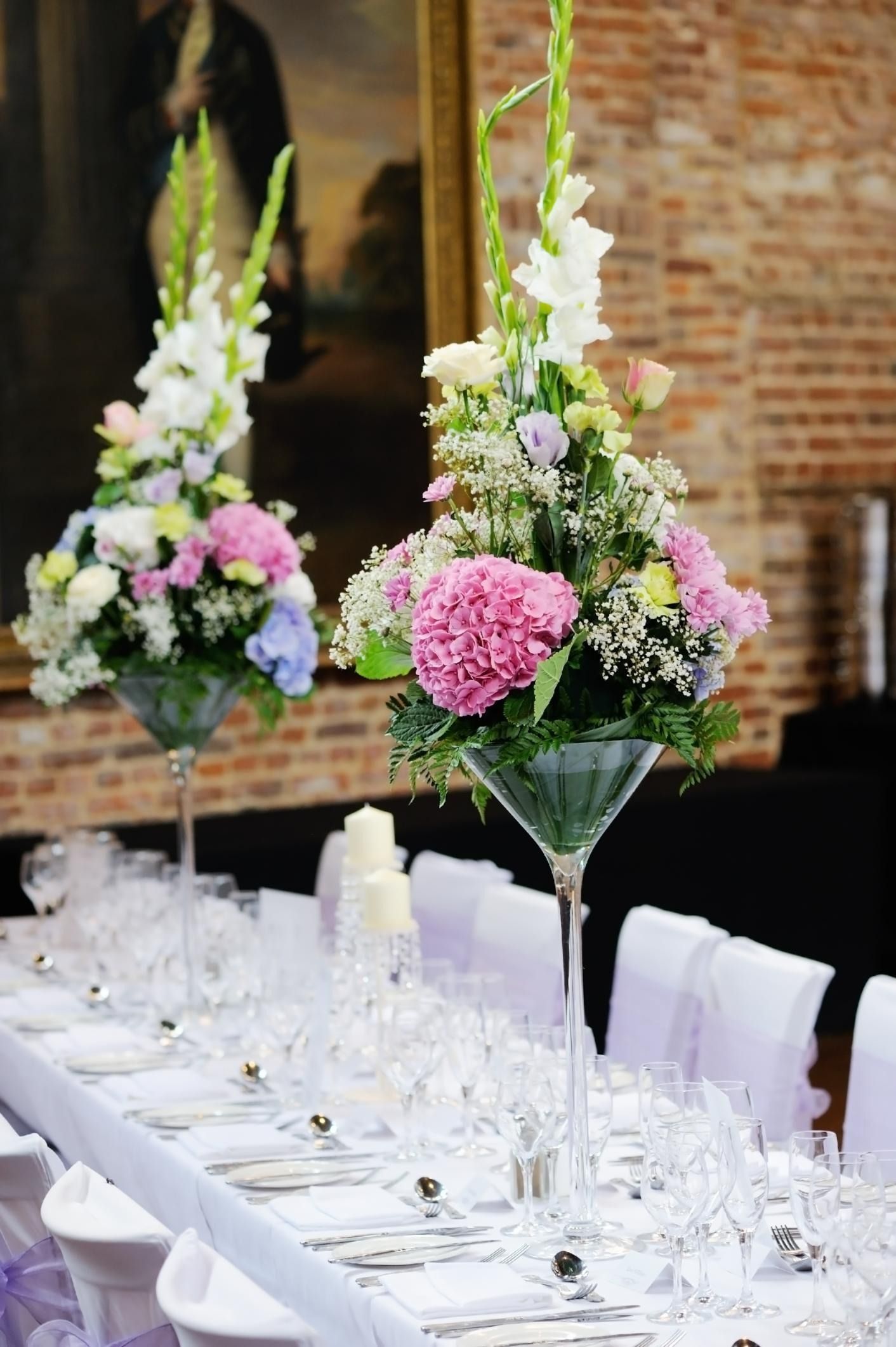 Elegant long banquet table with white place settings and tall pink-and-white floral centerpieces in a brick-walled venue