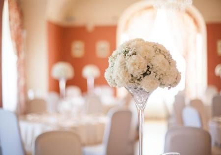 White floral centerpiece in a glass vase on a decorated banquet table with a softly blurred reception hall background
