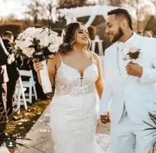 Newlywed couple in white wedding attire walking outdoors, smiling and holding hands by floral decorations