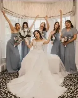 Bride seated with four bridesmaids in gray dresses, holding bouquets in a soft-lit room