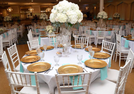 Elegant banquet table with white chairs, gold plates, and a large white floral centerpiece in a ballroom