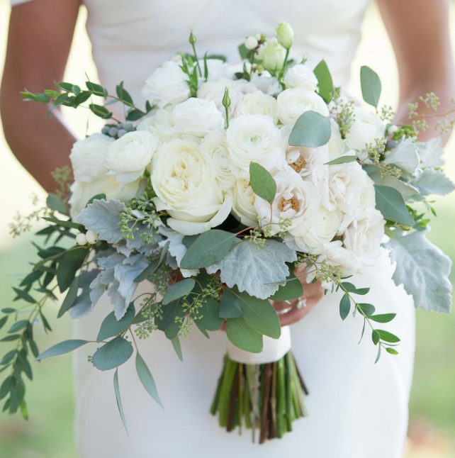 White bridal bouquet with roses, eucalyptus, and greenery held in front of a bride in a white dress