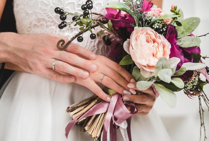 Hands holding a pink bridal bouquet with ribbon, against a white wedding dress.