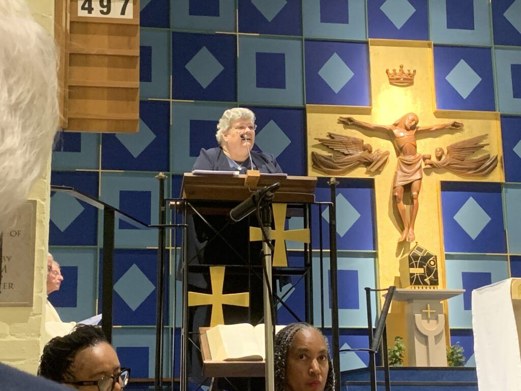 A woman stands at a podium in front of a cross with the number 497 on it