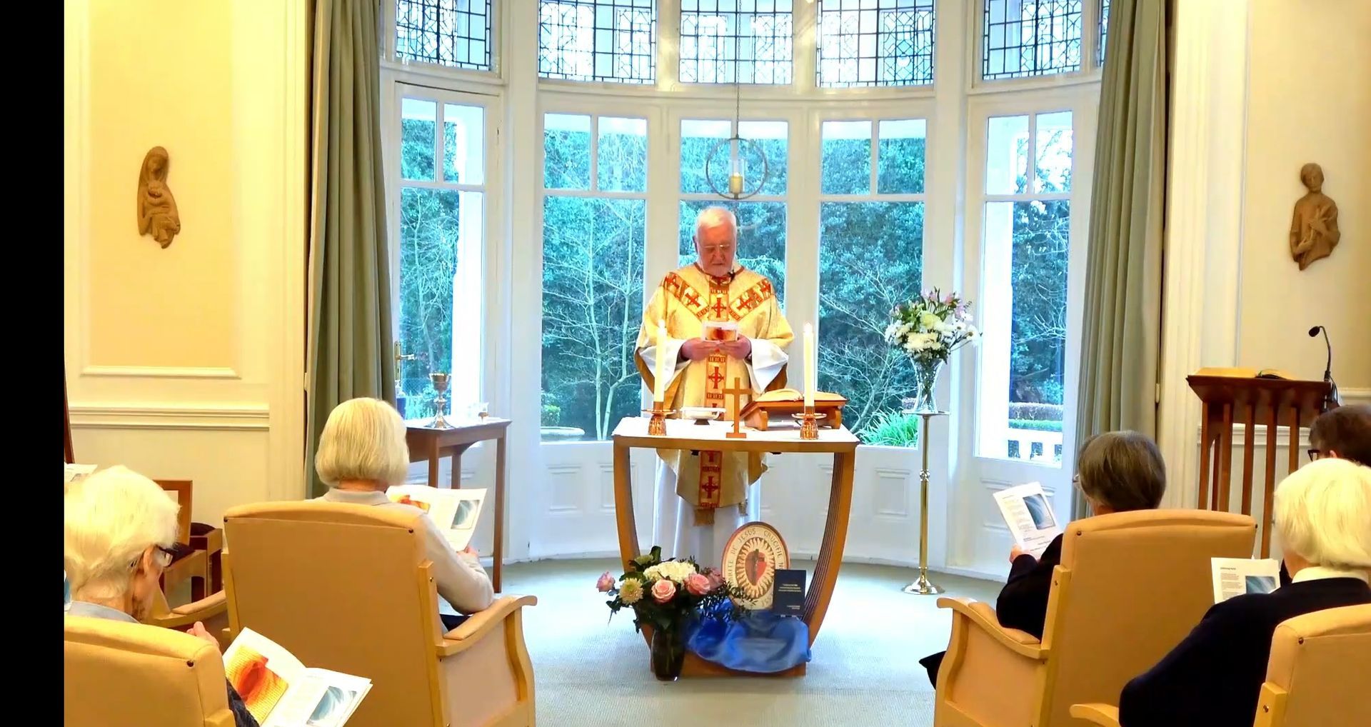 Priest in gold vestments leads a service in a room with a bay window.