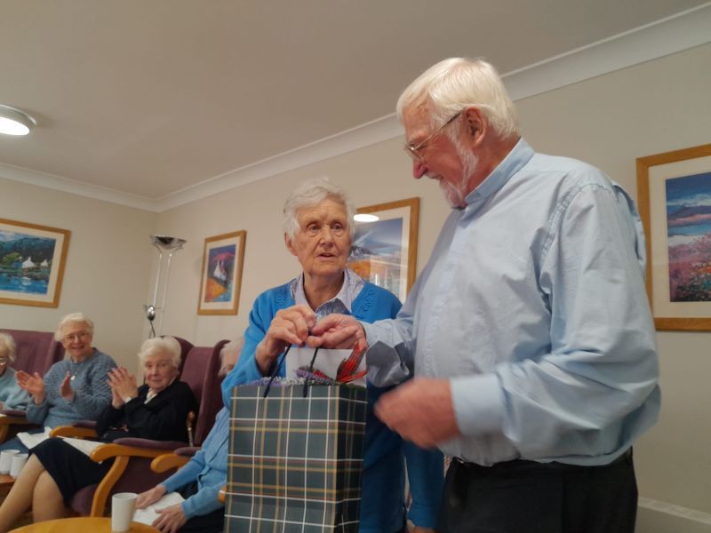 Elderly man gives gift to another elderly person in a room, while others clap.