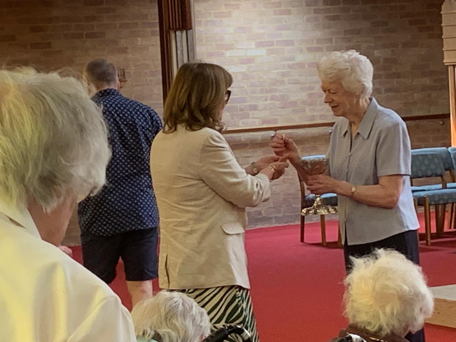 Woman handing something to another woman in a church setting. Red carpet, brick wall.