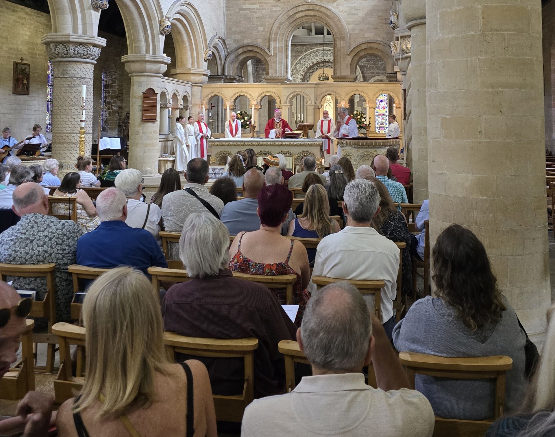 People seated in a church attend a service. Clergy stand at the altar; sunlight streams through windows.