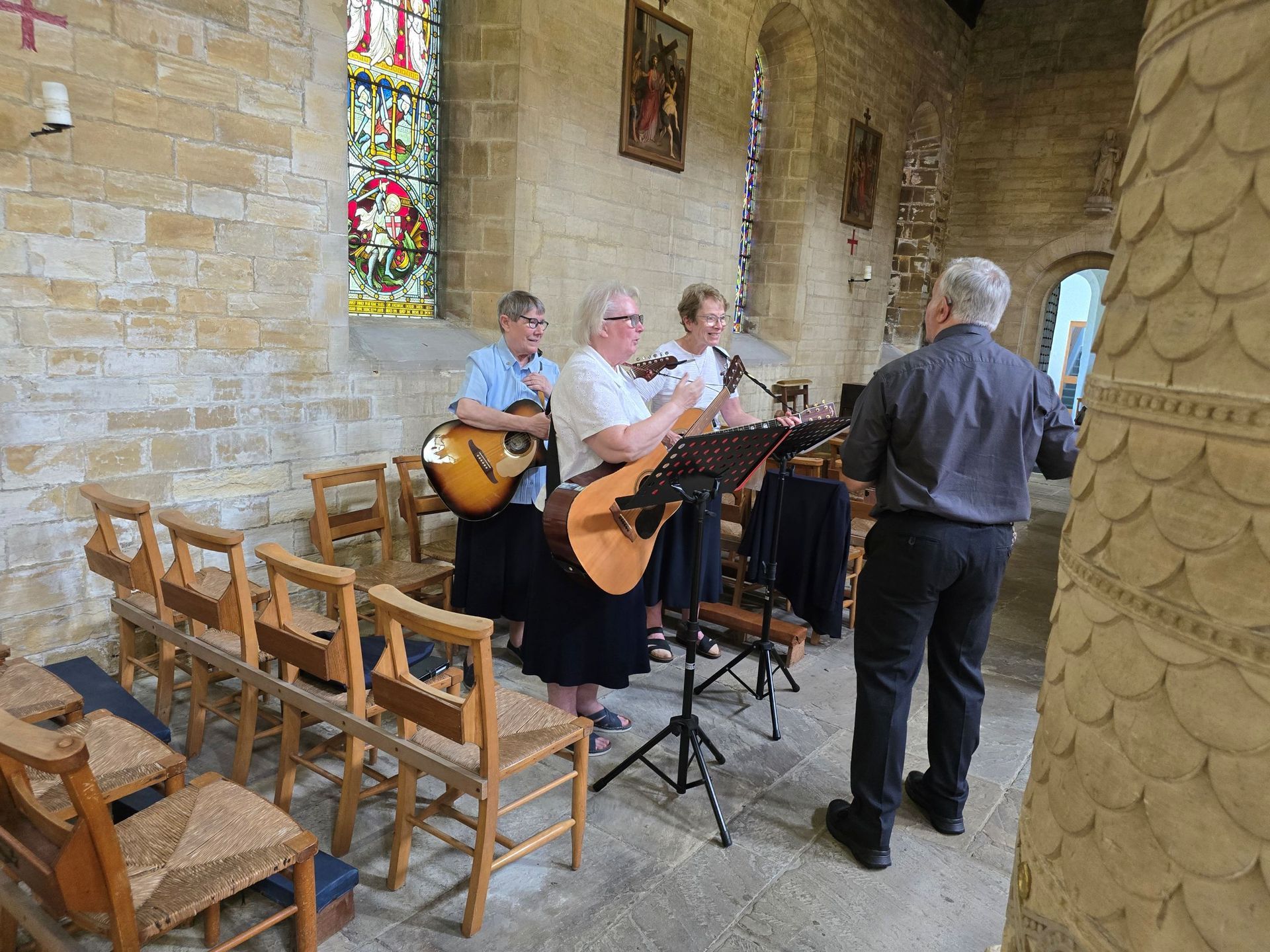Four musicians performing inside a stone church, using mandolin, guitar, and microphone. One man conducts.